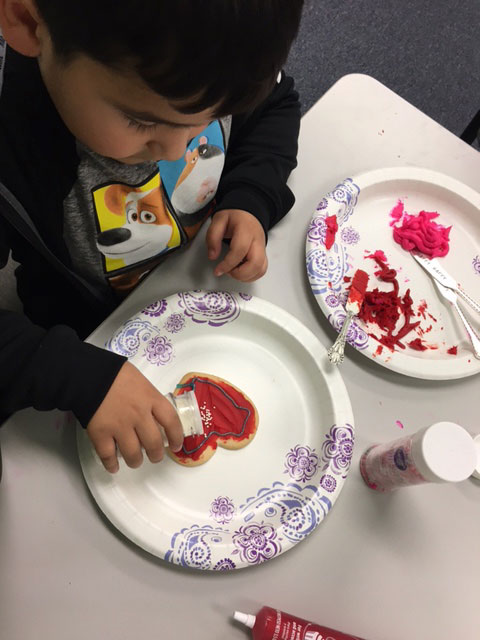 A child is decorating a heart shaped cookie with red sprinkles